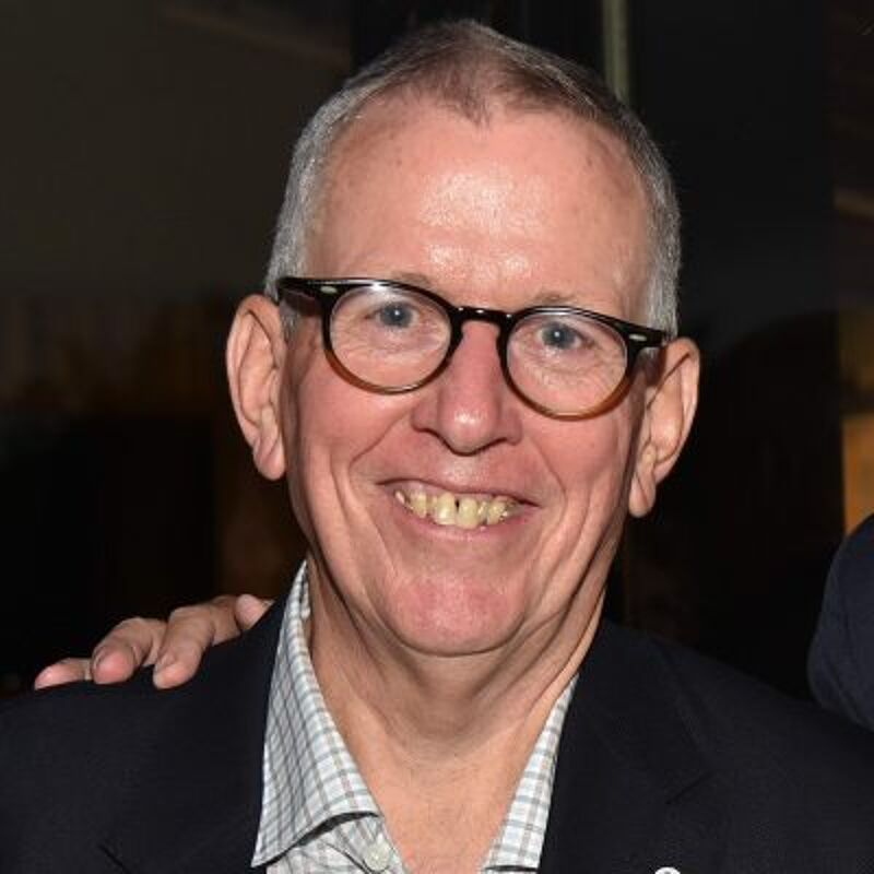 The image shows a man with glasses smiling at the camera. He has short gray hair and is wearing a collared shirt under a dark blazer. His teeth are visible, and he appears to be in a social setting. Someone's arm is on his shoulder.